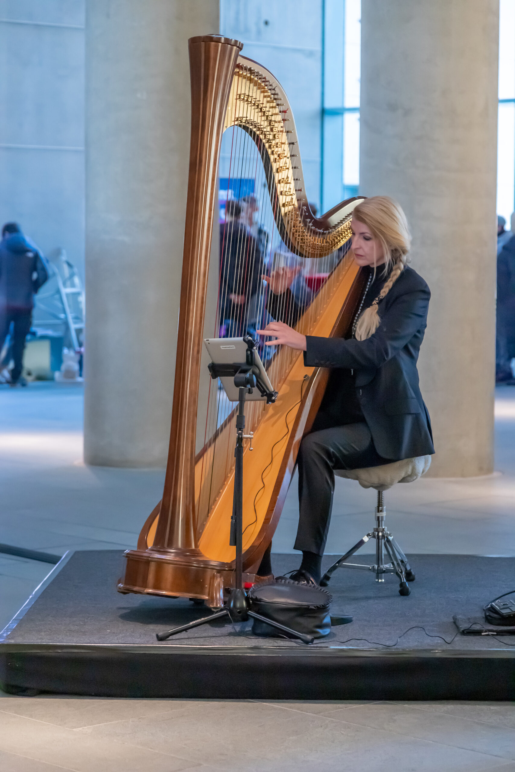 Person sitzt auf einem Hocker und spielt eine Harfe auf einer kleinen Bühne; neben dem Instrument ist ein Noten- oder Tabletständer, im Hintergrund ein heller Innenraum mit Säulen
