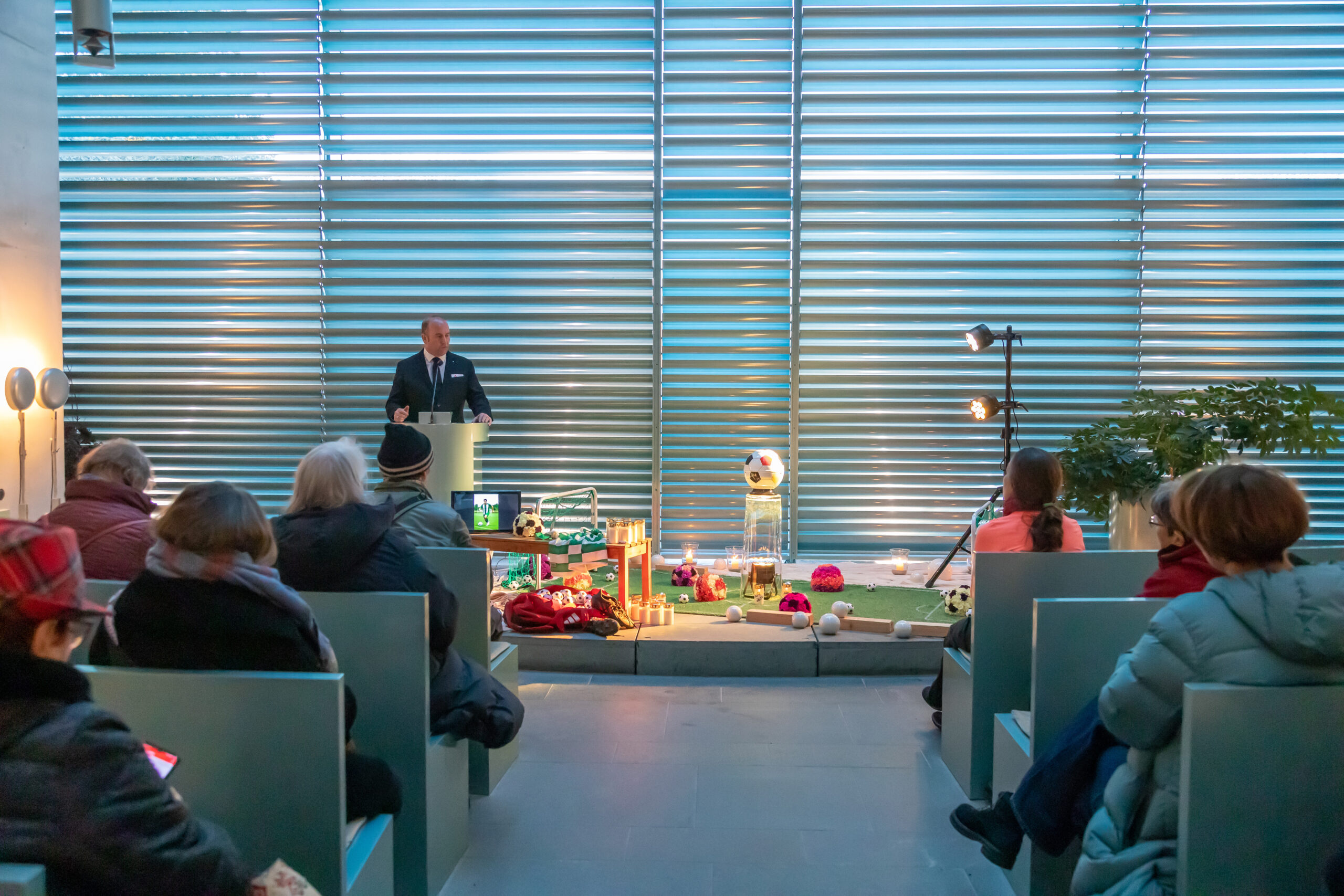 Person steht an einem Rednerpult und spricht vor sitzendem Publikum; vor der Bühne sind Kerzen, Blumen und persönliche Gegenstände dekoriert, in einem modernen Innenraum mit großen Fensterlamellen.