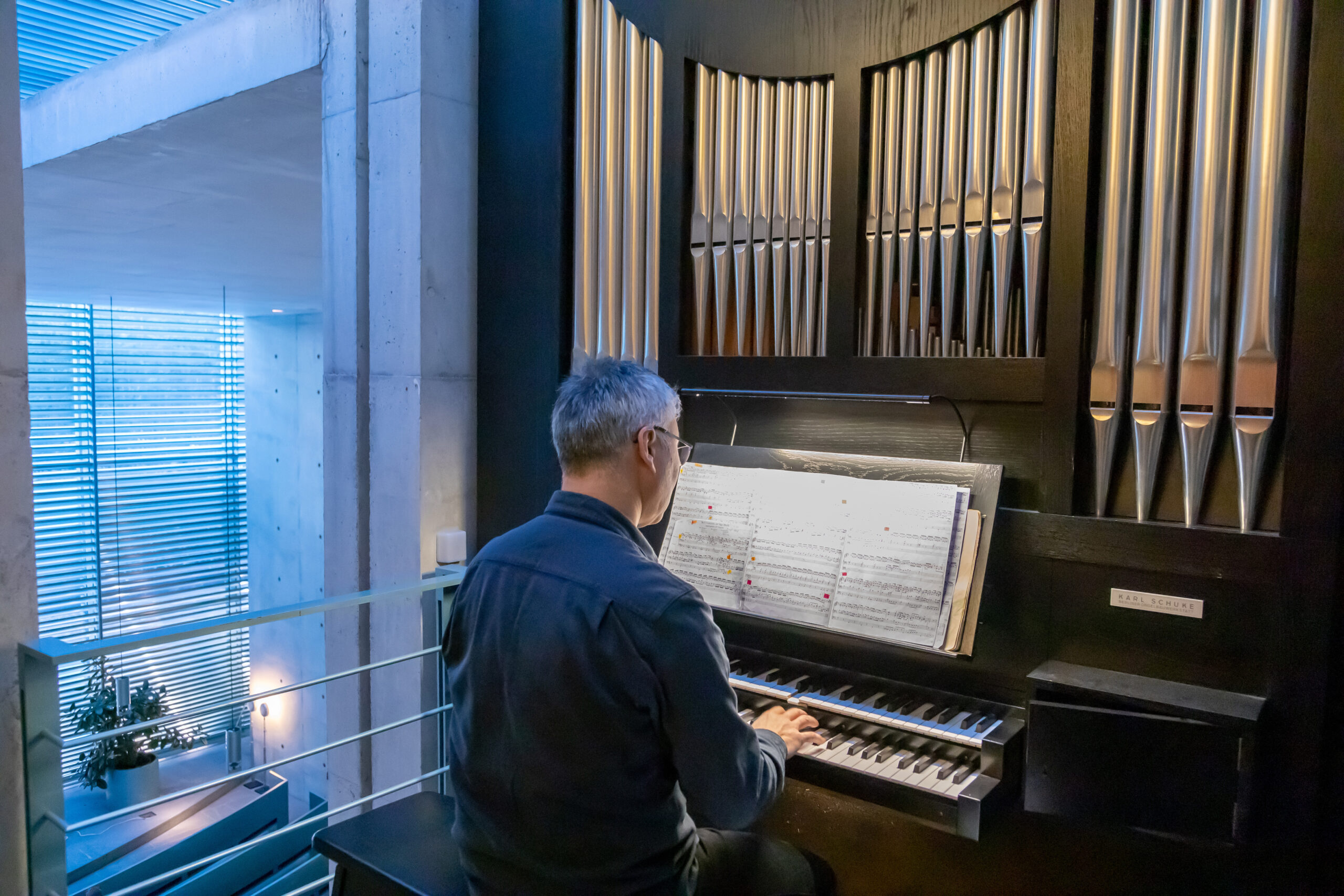 Person sitzt an einer Orgel und spielt nach Noten; im Hintergrund sind Orgelpfeifen und ein moderner Innenraum mit Betonwänden und Fensterlamellen zu sehen.